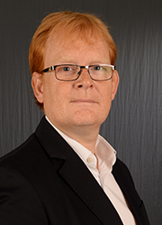 A man with reddish hair and glasses wearing a black blazer and white shirt against a dark background.