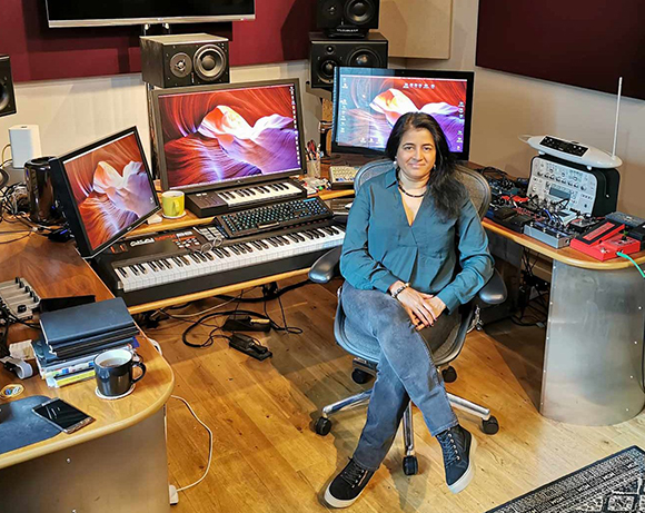 A woman sits in a chair amidst a music studio filled with monitors, keyboards, and audio equipment.