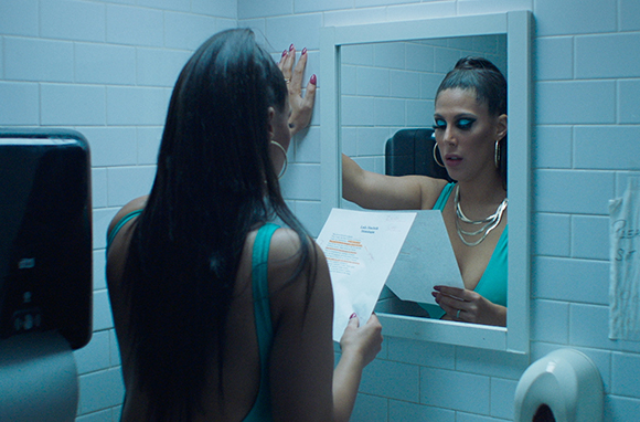 A woman with long dark hair looks into a mirror in a tiled bathroom while holding a white paper.