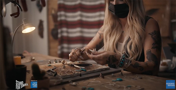 A person with blonde hair and tattoos crafts jewelry at a wooden workbench under a desk lamp.