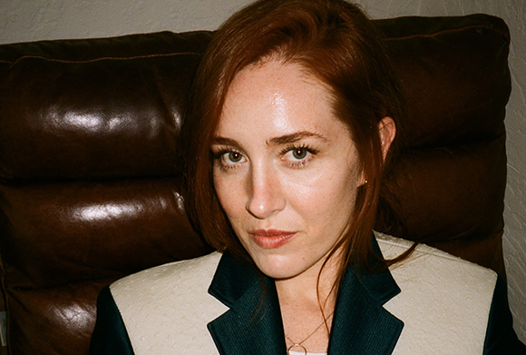 A woman with red hair and light eyes looks directly at the camera while sitting against a brown leather couch.