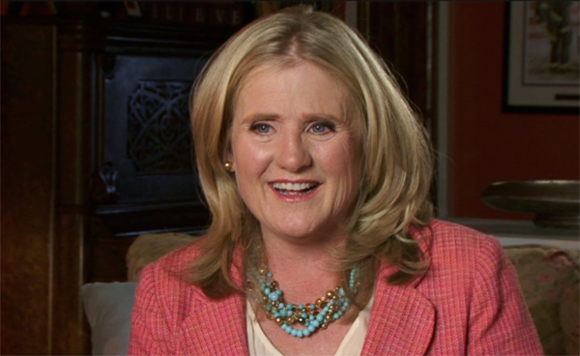 A smiling blonde woman in a pink blazer and turquoise beaded necklace.