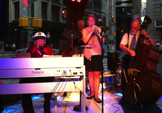 A jazz quartet performs on a lit stage outdoors, featuring a keyboardist, singer, and double bassist.