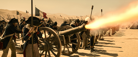 Soldiers in historical uniforms firing a large cannon that emits a bright orange flame in a desert setting.