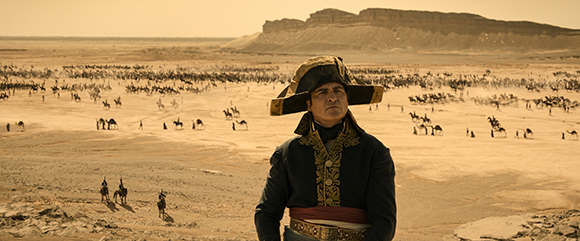Man in a dark blue military uniform and bicorne hat overlooks a desert plain filled with cavalry.