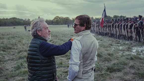 A man in a dark quilted vest gestures toward a man in a white shirt and beige vest in a grassy field.