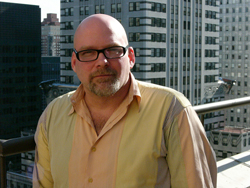A bald man with glasses and a goatee wearing a light yellow button-down shirt outdoors.