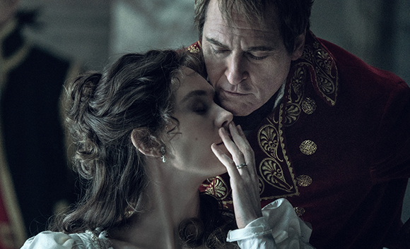 A man in a red embroidered jacket leans close to a woman with dark hair in a period setting.
