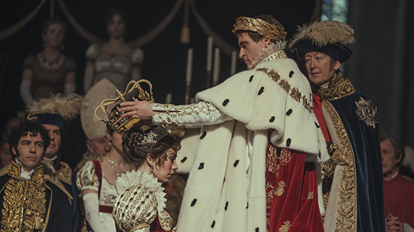 A man in a white ermine cloak and gold laurel wreath holds a crown over a kneeling woman in a period gown.