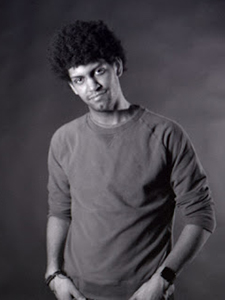 Black and white portrait of a man with curly hair wearing a long-sleeved shirt against a dark background.