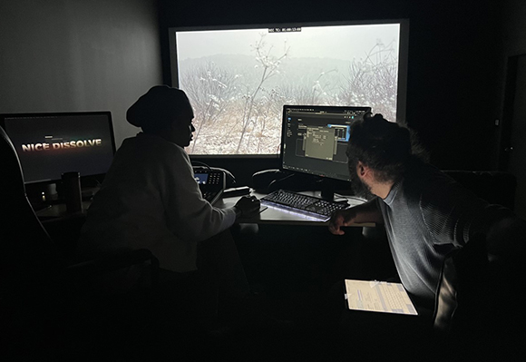 Two people sit in a dark room, silhouetted by the light of three computer monitors.
