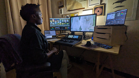 A person sits at a desk with four computer monitors displaying video editing software and footage.