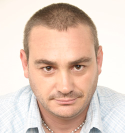 Close-up portrait of a man with short hair and light stubble wearing a light blue patterned shirt.