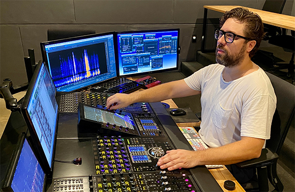 A man with glasses and a beard operates a large audio mixing desk with several computer monitors.