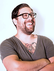 A smiling man with dark hair, glasses, and a beard wearing a grey v-neck t-shirt.