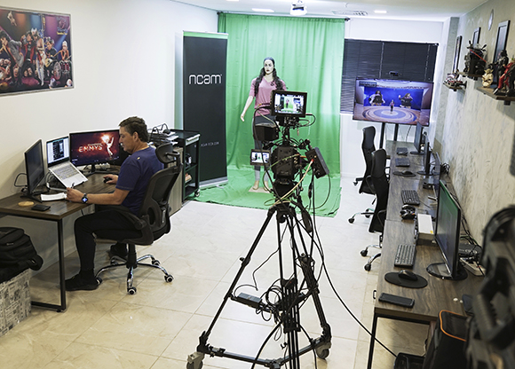 A man works at a desk in a studio featuring a green screen and a large camera on a tripod.