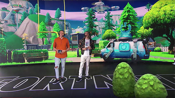Two men stand in a studio before a large backdrop featuring Fortnite landscapes and characters.