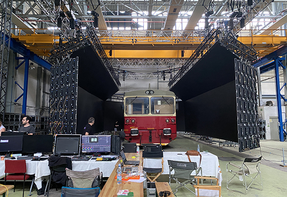A red train sits between two large black LED screens in a high-ceilinged industrial studio with production equipment.