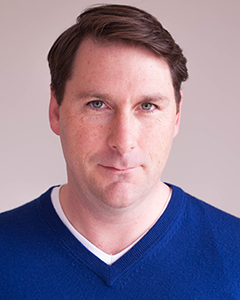 A man with short brown hair and blue eyes wearing a blue v-neck sweater against a plain background.