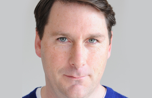 Close-up portrait of a man with light skin, brown hair, and blue eyes looking directly at the camera.