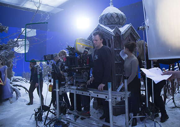 A film crew operates a camera in front of a miniature snowy building on a blue screen set.