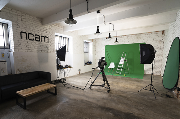 A photography studio featuring a green screen, camera on a tripod, softbox lights, and a black leather sofa.