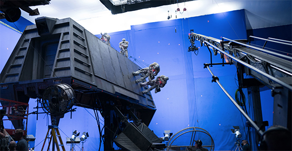 Actors in gear climb a large dark prop structure in front of a blue screen on a film set.