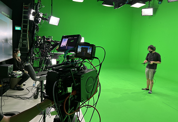 A camera crew films a person standing in a large green screen studio.