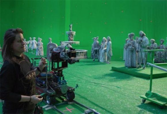 A woman operates a camera on a dolly in a green screen studio filled with various statues.