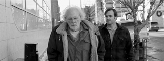 Black and white photo of an older man with white hair and a younger man walking outdoors on a city street.