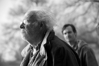 Black and white profile of an elderly man with glasses and white hair, with a blurred man in the background.