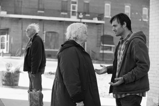 A man gestures while talking to an elderly woman on a sidewalk in front of a brick building.