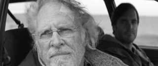 Black and white close-up of an older man with glasses and a beard sitting in a car.