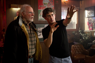 A man in a black t-shirt gestures with both hands while talking to an older man in a jacket.