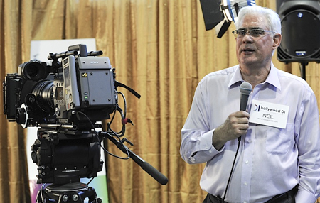 A man in a light blue shirt speaks into a microphone standing beside a large professional video camera.