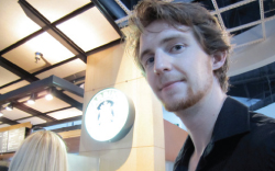 A young man with light brown hair and a goatee looking toward the camera in a brightly lit indoor space.