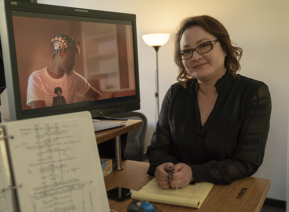 A woman in a black blouse sits at a desk next to a computer monitor displaying a video of a person.