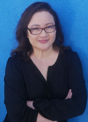 A woman with dark hair and glasses wearing a black shirt, posing with arms crossed against a blue wall.