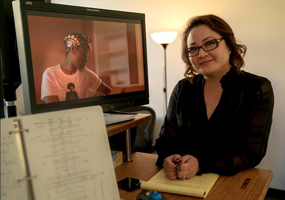 A woman with glasses smiling at the camera while sitting at a desk with a computer monitor and notepad.