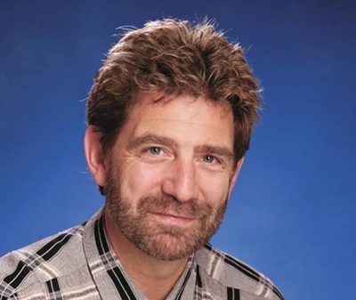 A man with wavy brown hair and a beard wearing a plaid shirt against a blue background.
