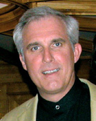 A man with gray hair and blue eyes smiling while wearing a black shirt and tan jacket.