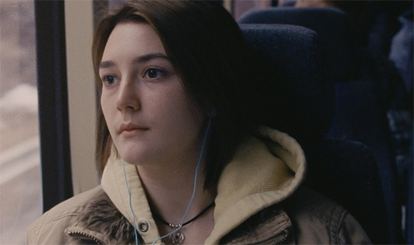 A young woman with dark hair sits on a bus, looking out the window while wearing blue earbuds.