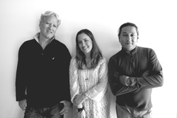 Black and white portrait of three smiling people standing together against a plain wall.