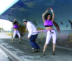 Two women in purple tops and white pants dance while a man in a white hoodie skateboards past them.