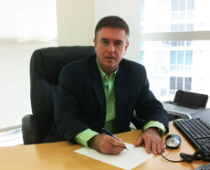 A man in a dark blazer and light green shirt sits at a desk holding a pen over a piece of paper.