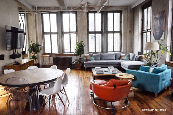 Industrial lounge with large windows, a grey sectional sofa, a blue armchair, and a round wooden table.