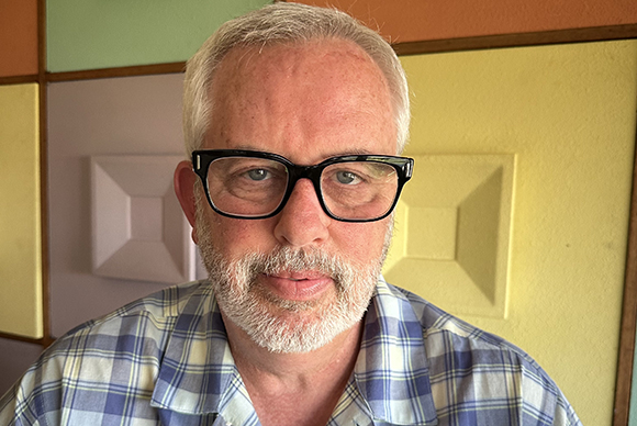 A man with short grey hair, black-rimmed glasses, and a grey beard wearing a blue plaid shirt.