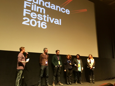 A group of five people stand on a stage in front of a large screen displaying "Sundance Film Festival 2016". One person on th