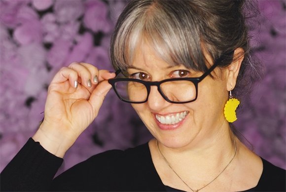A woman with grey hair and yellow earrings smiles while adjusting her black glasses against a purple background.