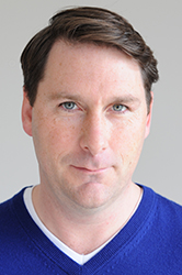 A man with short brown hair and light eyes wearing a blue v-neck sweater against a plain background.
